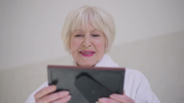 Portrait of happy smiling retiree looking at photo frame smiling in slow motion. Caucasian grey-haired beautiful woman recalling good memories standing at home indoors in bathrobe. — 비디오