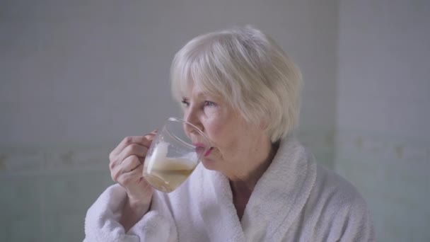 Hermosa mujer mayor con cabello gris bebiendo café en cámara lenta girando mirando a la cámara sonriendo. Retrato de feliz seguro relajado caucásico jubilado posando en el baño en casa con bebida caliente. — Vídeos de Stock