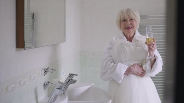 Gorgeous happy senior bride with wedding dress drinking champagne in slow motion smiling looking away. Portrait of attractive Caucasian woman standing in bathroom rejoicing holiday day. — Wideo stockowe