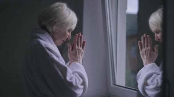 Side view reflection in mirror of sad senior Caucasian woman praying standing at window indoors. Upset depressed lonely retiree in bathrobe with hands together asking God for help and thanking. — стокове відео