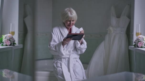 Wide shot crying senior beautiful woman sitting in bathroom with wedding dress looking at photo frame. Sad worried Caucasian lady with grey hair recalling memories reflecting in mirror. — Vídeo de Stock