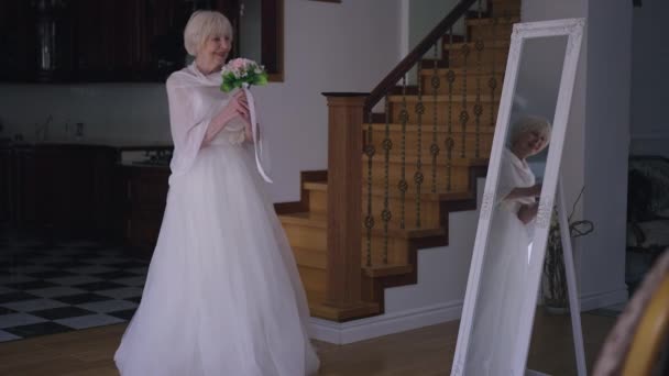 Wide shot excited happy senior bride in elegant wedding dress dancing with bridal bouquet in living room admiring reflection in mirror. Beautiful Caucasian woman with grey hair rejoicing marriage. — Stockvideo