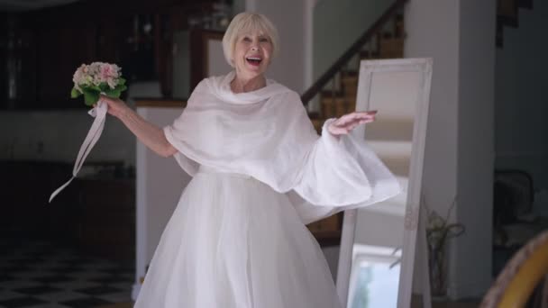 Joyful dance of happy elegant gorgeous senior woman in wedding dress with bridal bouquet indoors. Portrait of confident beautiful Caucasian woman with grey hair dancing smiling looking at camera. — Stockvideo