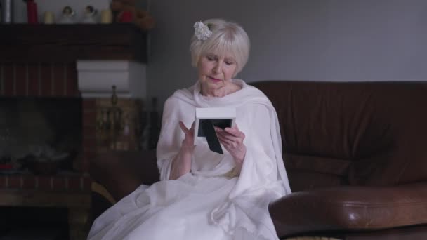 Thoughtful Caucasian widow in wedding dress sitting with photo in living room sighing looking away. Portrait of beautiful elegant bride recalling memories thinking in slow motion indoors. — Vídeo de Stock