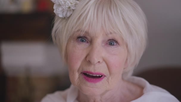 Headshot of gorgeous Caucasian senior woman in wedding dress with flower in hair smiling looking at camera. Close-up confident happy bride posing in slow motion indoors at home. Beauty and femininity. — Stock video