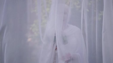 Happy senior woman in wedding dress passing white light curtain hanging outdoors looking at camera smiling. Portrait of confident Caucasian bride in wedding dress posing with bridal bouquet.