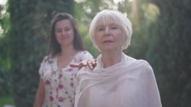 Beautiful senior elegant woman standing in sunrays in spring summer park with blurred granddaughter touching shoulder in slow motion. Portrait of happy retiree posing in sunlight outdoors.