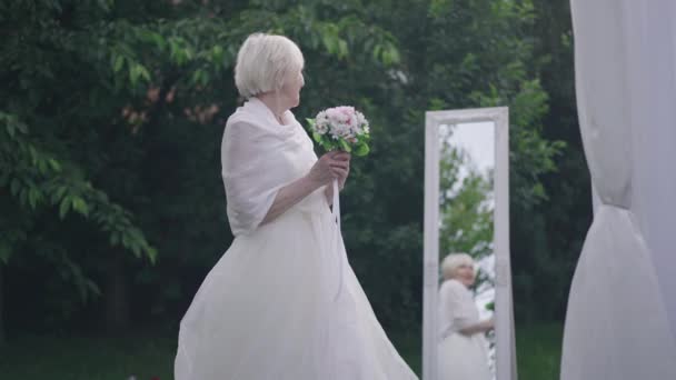 Side view happy senior bride throwing bridal bouquet in slow motion in spring summer park at white altar. Smiling satisfied Caucasian woman getting married outdoors. Traditions concept. — Stockvideo