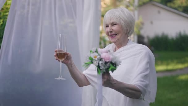 Satisfied beautiful senior woman in elegant wedding dress looking away listening compliments drinking champagne in slow motion. Portrait of happy Caucasian bride outdoors in summer park celebrating. — Video