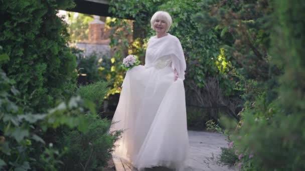 Wide shot gorgeous confident senior bride in white elegant wedding dress spinning in slow motion smiling looking at camera. Attractive Caucasian woman with grey hair posing in sunshine outdoors. — Stockvideo