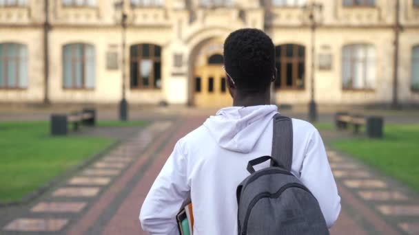 バックビューに自信を持って若い学生がゆっくりと屋外で大学のキャンパスに歩いてください。秋の日に大学に本と一緒に散歩インテリジェントアフリカ系アメリカ人男性。教育情報. — ストック動画