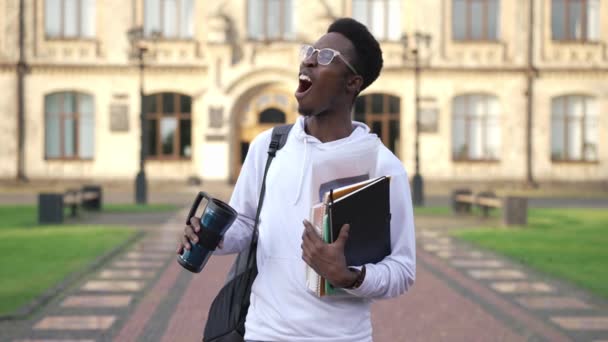 Tired African American man yawning standing at college campus with books and empty thermos. Portrait of sleepless exhausted young student outdoors in the morning. Education and lifestyle. — Stockvideo
