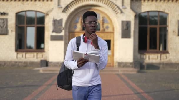 大学のキャンパスで読書本を読んで歩く思慮深いスマートなアフリカ系アメリカ人の若者。フロントビューポートレートのインテリジェント男性学生散歩で大学で午前中屋外. — ストック動画