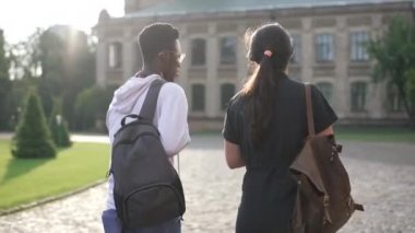 Back view young interracial couple of students walking on sunny morning to university campus outdoors. Confident smart African American man and Caucasian woman strolling talking in sunshine.
