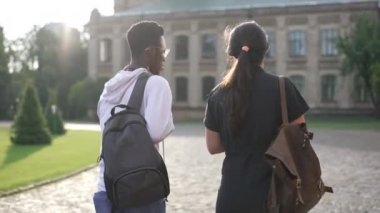 Back view young interracial couple of students walking on sunny morning to university campus outdoors. Confident smart African American man and Caucasian woman strolling talking in sunshine.