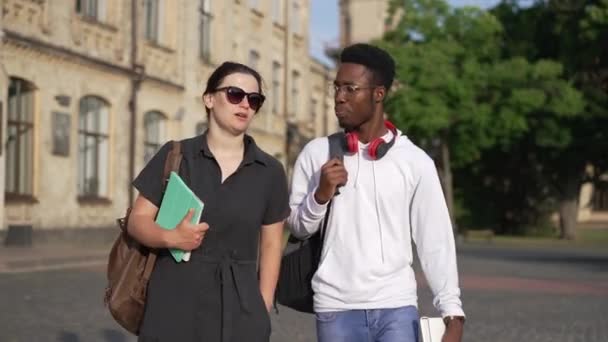 Dolly shot confident African American man and Caucasian woman walking outdoors talking at university campus. Interracial couple of groupmates discussing studies strolling on sunny morning at college. — Wideo stockowe