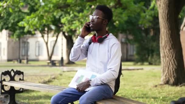 Positieve slimme Afro-Amerikaanse student in brillenglazen zittend op een bank en lachend aan de telefoon. Portret van een knappe intelligente jongeman in de ochtend op de binnenplaats van de universiteit. Onderwijs. — Stockvideo