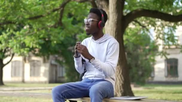 屋外の大学の庭でベンチに座って踊るヘッドフォンで楽しい若者の肖像画。アフリカ系アメリカ人の学生が午前中は趣味を楽しんでいます。教育と喜び. — ストック動画
