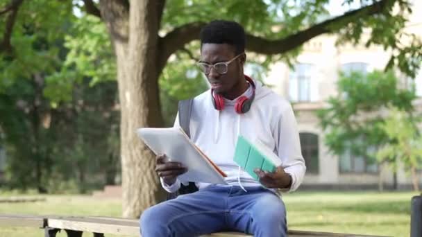 Zoom in to stressed overloaded young student sitting on bench with tablet and paperwork holding head in hands. Portrait of sad overworking African American man sighing studying at university yard. — стоковое видео