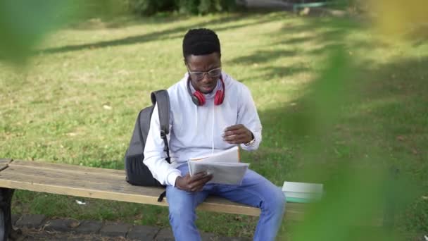 Yoğunlaşmış Afro-Amerikan öğrenci dışarıda üniversite bahçesinde bankta oturarak öğreniyor. Güneşli bir günde ders çalışmayı düşünen kendinden emin genç bir adamın portresi. Zeka ve eğitim. — Stok video