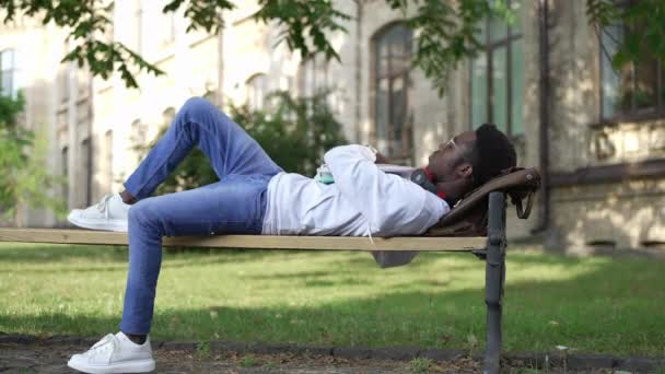 Estudiante afroamericano agotado durmiendo en el banquillo al aire libre despertándose apresurándose a salir corriendo. Joven hombre cansado inteligente corriendo con mochila y papeleo a los estudios. Educación y cansancio. — Vídeos de Stock