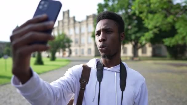 Portrait of young African American man taking selfie standing outdoors on university yard. Confident male student photographing at college campus. Lifestyle and education concept. — стокове відео