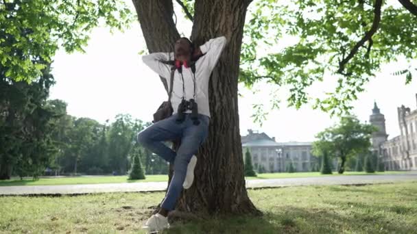 屋外の日差しの中で木に立ってリラックスしたアフリカ系アメリカ人の若い学生を撮影しました。大学の庭で自然の美しさを賞賛トランクにもたれて自信に満ちた幸せな男の肖像画. — ストック動画