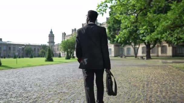 Tracking shot African American professor in suit walking on university yard in the morning. Back view confident intelligent young man strolling to college building outdoors. Education and profession. — Stok video