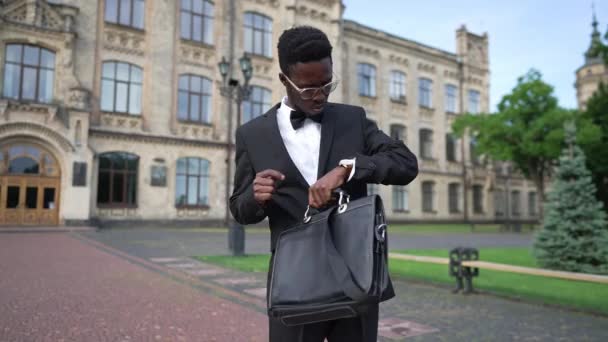 Medium shot portrait of self assured elegant African American man in suit with attache case standing at college campus outdoors talking gesturing. Happy new teacher at university in the morning. — стоковое видео