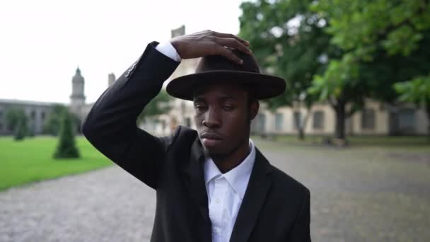 Elegant African American man putting on stylish hat dancing singing outdoors. Portrait of confident handsome young student in suit having fun posing at university campus on spring autumn day. — Video