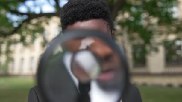 Close-up portrait of African American genius scientist posing with magnifying glass outdoors. Curios young serious man looking at camera lifting lowering magnifier. Science and lifestyle concept. — стокове відео