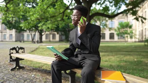 Overburdened African American young man in suit sitting on bench talking on phone and on tablet. Portrait of busy overworking businessman discussing ideas outdoors. Multitasking concept. — Wideo stockowe