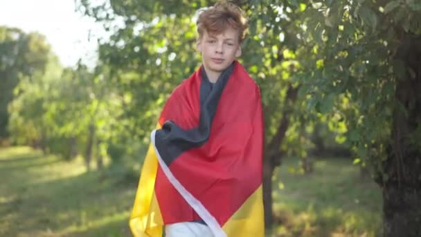 カメラの笑顔を見てドイツの旗に包まれた巻き毛を持つ10代の白人の赤毛の少年の肖像画。公園で晴れた春の夏の日にポーズをとる幸せなリラックスしたティーンエイジャー。プライドとライフスタイル. — ストック動画