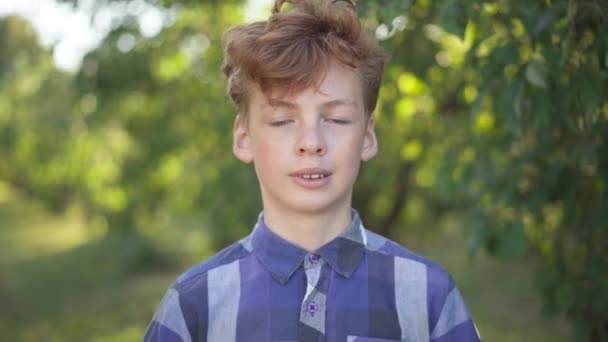 Close-up portrait of confident Caucasian teenage boy with red curly hair smiling looking at camera standing in summer spring park. happy carefree adolescent teenager posing outdoors. — Stok video