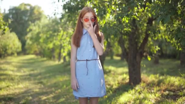 Zoom in to happy Caucasian teenage redhead girl in sunglasses standing in sunshine smiling looking at camera. Portrait of carefree confident teenager with red long hair posing in sunlight in park. — Wideo stockowe