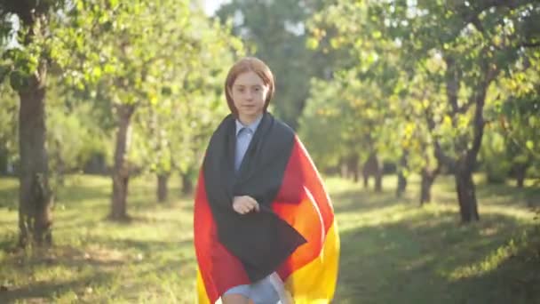 Confident proud teenage redhead girl wrapping in German flag walking in sunrays looking at camera. Portrait of pretty Caucasian teenager posing strolling in spring summer park outdoors. Dolly shot. — Vídeo de Stock