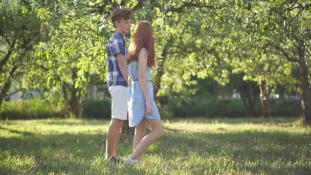 Side view wide shot happy loving teenage couple holding hands running in sunshine outdoors. Tracking shot Caucasian redhead boy and girl having fun dating in spring summer park. First love and joy. — Video