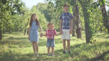 Three redhead children holding hands walking to camera leaving in sunshine outdoors. Happy little boy strolling in spring summer park with teenage brother and sister. Weekend leisure and family.