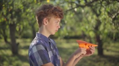 Side view handsome teenage boy with red hair launching paper plane outdoors looking away. Portrait of confident Caucasian adolescent teenager playing in spring summer park having fun on weekend.