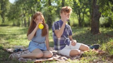 Wide shot two teenagers blowing balloons sitting in sunrays on summer spring meadow in park. Relaxed Caucasian teenage boy and girl enjoying weekend leisure outdoors in sunshine.