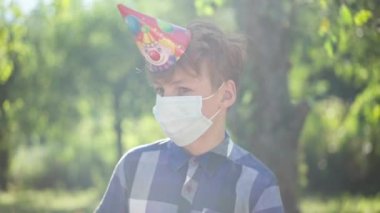 Sad Caucasian teenage redhead boy in coronavirus face mask and party hat in sunrays outdoors alone on birthday. Portrait of upset teenager in summer spring park on lonely picnic. Covid-19 new normal.