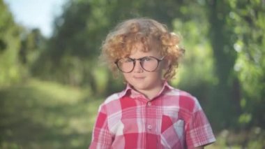 Nerd cute redhead little boy in eyeglasses posing in sunny spring summer park outdoors. Portrait of confident smart Caucasian happy kid looking at camera smiling. Intelligence and leisure.