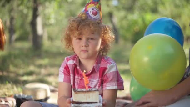 Portrait of charming little redhead Caucasian boy in party hat blowing candle on birthday cake smiling looking at camera. Happy child posing in sunshine in park celebrating with unrecognizable friends — стоковое видео