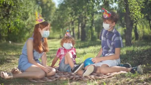 Wide shot happy Caucasian siblings sitting on spring summer picnic in park talking. Relaxed redhead boys and girl in party hats and coronavirus face masks celebrating birthday outdoors in sunshine. — ストック動画