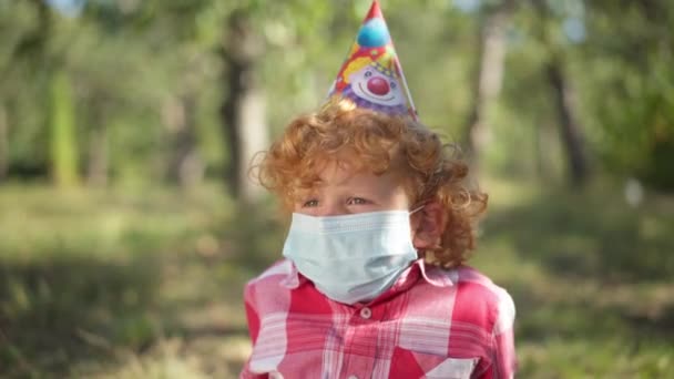 Cute Caucasian redhead little boy in Covid-19 face mask and party hat looking away sitting in sunshine in spring summer park. Lonely sad child outdoors alone on birthday on coronavirus pandemic. — Stockvideo