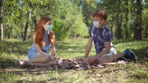 Teenage Caucasian couple in coronavirus face masks sitting in spring summer park talking. Wide shot portrait of redhead boy and girl dating outdoors on Covid-19 pandemic having picnic. — стоковое видео