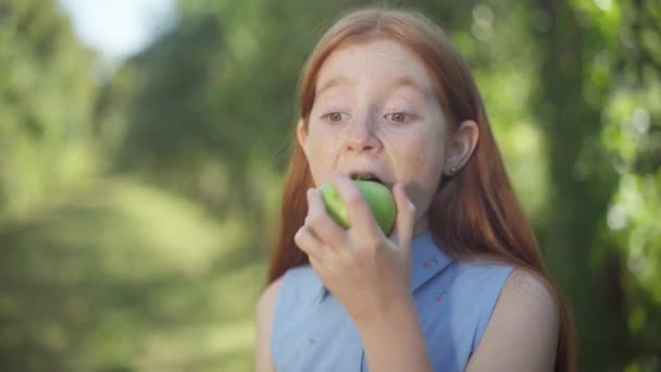 Confident charming redhead teenage girl biting chewing healthful apple smiling looking at camera. Portrait of happy Caucasian adolescent teenager eating fruit enjoying taste posing in park outdoors. — 图库视频影像