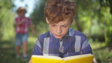 Rack focus from concentrated teenage boy reading book in sunny park to cheerful little brother running from background covering eyes with hands playing with sibling. Happy brothers resting outdoors.