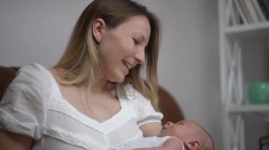 Happy loving Caucasian mother talking to newborn son kissing infant nose in slow motion. Portrait of smiling beautiful woman taking care of child indoors at home in living room. Love and maternity.
