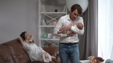 Portrait of happy father with baby son in hands standing in living room as relaxed mother listening to music in headphones sitting on couch. Loving Caucasian man taking care of newborn infant.